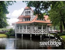 Ein Fachwerkgebäude mit roten Dachziegeln, das auf Stelzen über einem Teich errichtet ist, umgeben von Bäumen und grüner Landschaft. Rechts führt eine Brücke zum Gebäude, das als Restaurant, Hotel und Seebrücke dient.