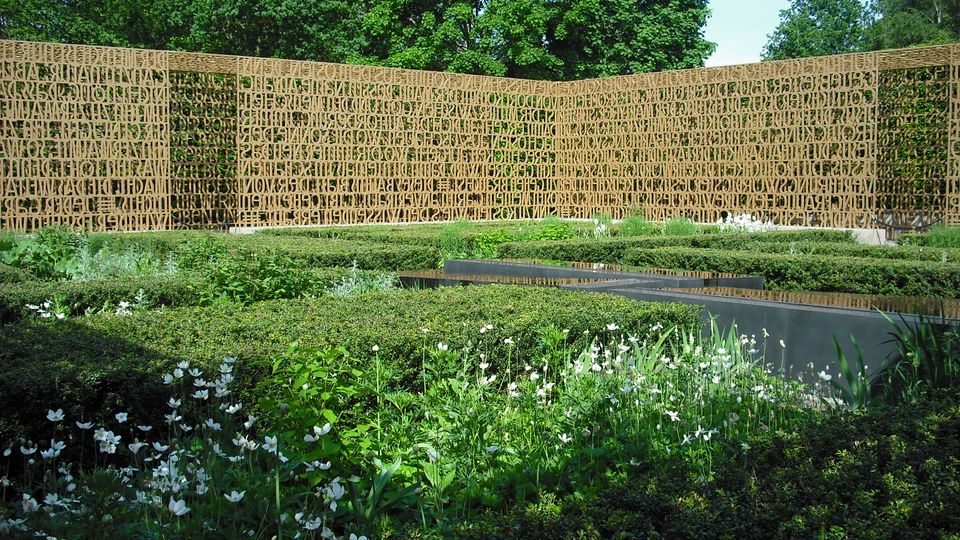 Gartenanlage mit niedrigen Hecken und Beeten voller weißer Blüten, in der Mitte ein rechteckiges Wasserbecken mit dunkler Einfassung. Dahinter steht eine lange, gebogene Sichtwand aus goldfarbenem Metallgitter mit ausgeschnittenen Buchstaben, umgeben von hohen Bäumen.