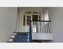 Treppenhaus mit blauem Teppichbelag auf den Stufen, weißem Holzgeländer und einer Doppeltür mit Glaseinsätzen und Rundbogen im oberen Bereich. Rechts befindet sich ein Fenster, das Tageslicht hereinlässt.