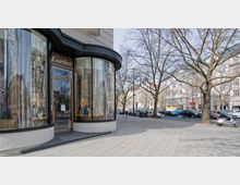 Ecke eines eleganten Geschäfts mit großen, gewölbten Glasfenstern und der Aufschrift „Cartier“ über der Eingangstür. Im Hintergrund eine breite Straße mit geparkten Autos, Bäumen ohne Laub und mehrstöckigen Wohngebäuden.