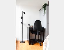 Ein schlichtes Homeoffice-Setup in einer Ecke eines hellen Raums mit einem schwarzen Schreibtisch, einem drehbaren Bürostuhl, einer schlanken Stehlampe und einer Zimmerpflanze, die von einem grauen Schrank herunterhängt. Der Boden ist aus Parkett, die Wände sind weiß.