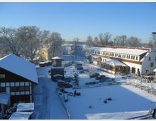 Historische Dorfansicht im Winter mit Schnee bedeckt, inklusive Fachwerkgebäuden, einem zentralen Platz mit parkenden Autos und einem kleinen Turm. Im Hintergrund sind Bäume und weitere Gebäude unter klarem, blauem Himmel zu sehen.