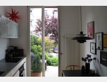 Moderne Küche mit weißen Schränken, einer grauen Arbeitsplatte und einem Herd, daneben eine Glas-Balkontür, die zu einer begrünten Terrasse mit Pflanzen und Möbeln führt. Rechts hängt eine schwarze Lampe über einem Esstisch mit Vasen und Bildern an der Wand.