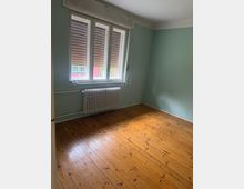 Leeres Zimmer mit hellblauen Wänden und holzfarbenem Dielenboden, beleuchtet durch ein großes dreiteiliges Fenster. Unter dem Fenster befindet sich ein weißer Heizkörper; außen sind Rollläden teilweise heruntergelassen.