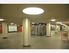 Unterirdischer Gehweg mit mehreren großen, runden Metallpfeilern, leuchtenden Deckenlampen, einem gläsernen Aufzug und bunten Wandverkleidungen in Rot, Gelb und Weiß. Im Vordergrund steht ein Werbeschild für Backwaren.