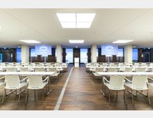 Großer Konferenzraum mit weißen Stühlen und Tischen, ausgerichtet auf ein Rednerpult in der Mitte. Der Raum hat einen Holzboden, helle Deckenbeleuchtung und an der Vorderseite eine Leinwand mit dem Schriftzug "Olympiastadion" und einem Logo.