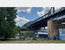 Eine erhöhte Eisenbahnbrücke aus Metall, gestützt von Stahlträgern, mit umliegenden Bäumen, graffitibesprühten Wänden und einem Kleinbau mit der Aufschrift „Hier wird gebacken“. Der Himmel ist leicht bewölkt, und im Vordergrund befindet sich eine kleine Grünfläche.