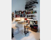 Ein kleiner Arbeitsbereich in einer Ecke eines Zimmers mit weißen Wänden und hellen Holzböden. An der Wand befinden sich mehrere weiße Regalbretter voller Bücher, Aufbewahrungsboxen und diverser Utensilien; darunter ein kleiner Tisch mit Metallgestell, ergänzt durch einen Hocker mit rundem Sitz.