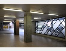 Unterführung mit beleuchtetem Flur, Fliesenboden und gitterförmigen Fensterelementen aus geometrischen Mustern. Im Hintergrund ist ein Eingang zum U-Bahnhof "Bundesplatz" und eine Straßenszene erkennbar.