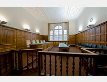 Innenansicht eines Gerichtssaals mit Holzvertäfelung an den Wänden, einem dekorativen Deckenmuster und einem großen Fenster im Hintergrund. Zu sehen sind ein Richterpult, mehrere Tische mit Stühlen und eine Trennung durch Holzgeländer.