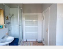 Helles Badezimmer mit weiß gefliesten Wänden, Waschbecken links und einer verglasten Duschkabine. An der Stirnwand steht ein großer weißer Handtuchheizkörper, rechts befindet sich eine weiße Tür; der Boden ist gefliest und mit einem kleinen Badvorleger versehen.