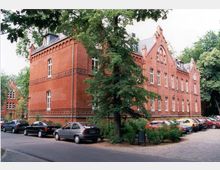 Rotes Backsteingebäude im neugotischen Stil mit dekorativen Fassadenelementen, spitzen Giebeln und weißen Fensterrahmen, umgeben von Bäumen und einer Reihe parkender Autos entlang einer Straße.