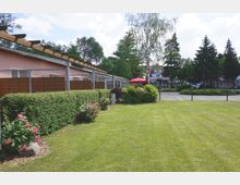 Rasenfläche mit Rosensträuchern und Hecke im Vordergrund, entlang einer niedrigen Gebäudereihe mit Holzpergola. Im Hintergrund sind Bäume, ein Parkplatz und Gebäude mit weißen Fassaden zu sehen, einschließlich eines Sonnenschirms in Rot.