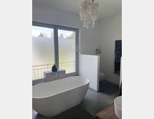 Modernes Badezimmer mit freistehender, ovaler Badewanne vor einer großen Fensterfront mit Milchglas. An der Decke hängt ein dekorativer Kronleuchter, und im Hintergrund sind eine teilabgetrennte Toilette sowie minimalistische Dekoration zu sehen.