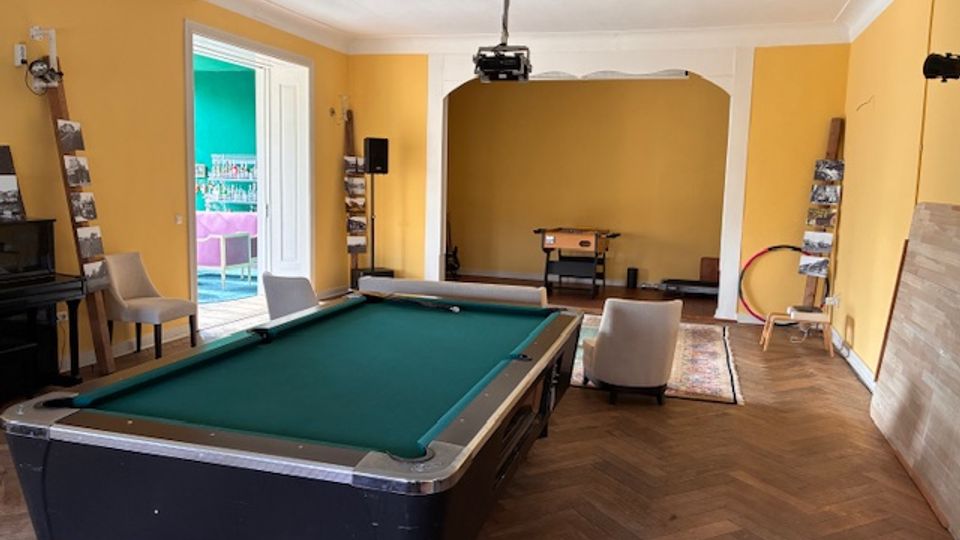 Großer Innenraum mit gelb gestrichenen Wänden, Holzboden und einem Billardtisch im Vordergrund. Hinten befindet sich eine breite Durchgangsöffnung mit einem hängenden Deckenprojektor, daneben Stühle, Regale und eine offene Tür zu einem angrenzenden, farbig gestrichenen Raum.