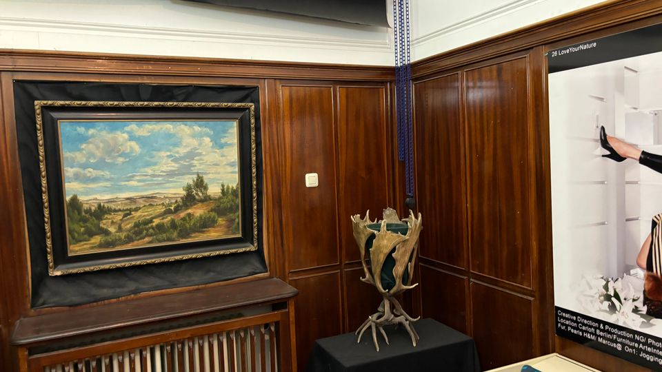 Innenraum mit dunkler Holzvertäfelung und weißer Decke, in dem ein gerahmtes Landschaftsgemälde über einem Heizkörper hängt. Daneben steht auf einem schwarz bedeckten Tisch eine skulpturale Geweih-Installation, rechts ist ein großes Wandposter zu sehen.