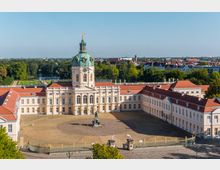 Luftaufnahme des Schlosses Charlottenburg in Berlin, mit einer barocken Fassade, zentralem Kuppelturm, einem Vorplatz mit Reiterdenkmal und einer goldenen Eingangspforte. Im Hintergrund ist ein Park mit einem Wasserlauf sowie umliegende Gebäude der Stadt sichtbar.