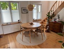 Heller Essbereich mit rundem Holztisch und vier Stühlen auf einem runden Teppich, daneben eine Holztreppe mit Geländer. Große Fenster mit halb geschlossenen Plissees, eine geflochtene Hängelampe sowie eine Kommode mit Pflanzen stehen an den Wänden.