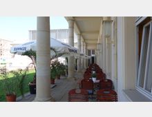 Außenterrasse eines Restaurants mit Holzstühlen und Tischen in einer überdachten Arkade mit steinernen Säulen. Über den Sitzbereichen spenden weiße Sonnenschirme mit der Aufschrift „Erdinger“ Schatten, und entlang der Terrasse stehen zahlreiche Blumentöpfe mit grünen Pflanzen.