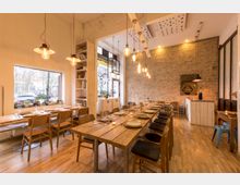 Ein schick eingerichteter Speiseraum mit Holzboden, langen Holztischen und passenden Stühlen. Der Raum ist von warmem Licht beleuchtet, hat freiliegende Ziegelwände, große Fenster mit Blick nach draußen und moderne Hängelampen.