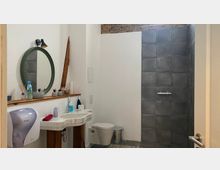 Ein modernes Badezimmer mit weißen Wänden, einer offenen Dusche mit dunkelgrauen Fliesen, einem runden Wandspiegel, einem Waschbecken und einer Toilette. Oben ist eine freiliegende Backsteinmauer sichtbar, und die Einrichtung beinhaltet minimalistische Dekorationen und Hygieneartikel.