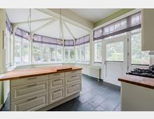 Helle Küche mit großem Kücheninselblock aus Holz und hellen Schrankfronten, dunklem Fliesenboden und Gasherd. Rundumlaufender Wintergarten-Erker mit vielen Sprossenfenstern, sichtbaren Deckenstreben, zwei Hängelampen sowie Tür nach außen.