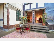 Eingangsbereich eines Gebäudes mit blau-weiß gekachelter und verputzter Fassade, großen Fenstern und einer offenen Tür zu einem Innenraum mit Kronleuchtern. Davor stehen auf einem gemusterten Teppich ein runder Tisch mit zwei roten Polsterstühlen, daneben Blumendekor, Topfpflanze und eine kleine Treppe zum Eingang.