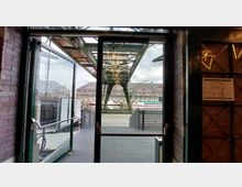 Blick durch eine Glastür in Richtung eines Kanals mit einem grünen Stahlgerüst oben, dahinter Gebäude im Backsteinstil und geparkte Boote. Die Umgebung wirkt industriell geprägt, mit Hinweisen auf eine Hafennähe.