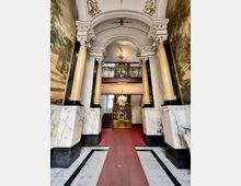 Prunkvoller Eingang eines historischen Gebäudes mit hohen Marmorsäulen, verziertem Stuck an der Decke und Wandgemälden auf beiden Seiten. Im Hintergrund führen eine Treppe und ein kunstvoll verzierter Holzaufzug zu den oberen Etagen.