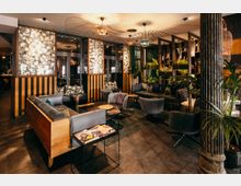 Gemütlicher Lounge- und Barbereich mit dunklen Sofas und Sesseln um niedrige Beistelltische, warm beleuchtet durch mehrere Hängelampen. Im Hintergrund stehen Pflanzenregale und Trennwände mit floral gemusterten Paneelen vor großen Glasfenstern und Türen.