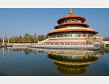 Runder, mehrstöckiger chinesischer Tempel mit geschwungenem, gelbem Dach, verziert mit roten und grünen Elementen, gelegen an einem künstlichen Teich, der das Gebäude spiegelt. Im Hintergrund sind Bäume und ein klarer blauer Himmel sichtbar.
