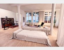 Offener Wohnbereich mit hellem Holzboden, großen Fensterfronten und durch Vorhänge teilweise verdecktem Tageslicht. Der Raum ist mit einem hellen Sofa, einem schwarzen Liegestuhl, Regalen, Teppichen und Pflanzen eingerichtet.