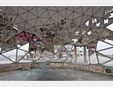 Inneres einer verlassenen Radarkuppel mit geometrischem Kuppeldach aus Dreiecksstrukturen, das mit Graffiti und Zeichnungen bedeckt ist. Der Blick nach draußen zeigt eine teilweise zerstörte Plattform und eine graue, neblige Landschaft im Hintergrund.