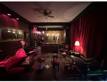 Ein schummrig beleuchteter Raum im Stil eines Vintage-Lounges mit roten Samtmöbeln, dunklen Holzböden und roten Vorhängen. Im Hintergrund befindet sich eine Bar mit Holztheke und Flaschenregalen, darüber hängen traditionelle Lampen.