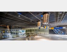 Modernes unterirdisches Bahnhofsinterieur mit glänzenden Metallpaneelen an der Decke, Rolltreppen, Glasgeländern und blau-gelben Wegweisern. Im Hintergrund sind ein Fahrgastbereich, ein Treppenaufgang und große Lichtinstallationen sichtbar.