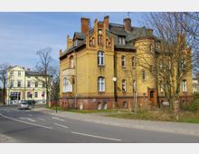 Mehrstöckiges Gebäude aus gelbem und rotem Klinker mit detailreichen Zierelementen, umgeben von kahlen Bäumen, neben einer Straße. Im Hintergrund steht ein helles Wohn- oder Geschäftshaus.