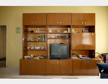 Holzvertäfelte Wohnwand im Retro-Stil mit offenen und geschlossenen Schränken, darin Geschirr, Gläser, Bücher und Dekorationsgegenstände. In der Mitte befindet sich ein Röhrenfernseher, daneben weitere Alltagsgegenstände auf der Ablagefläche; im Hintergrund gelbe Wände und ein Durchgang mit Vorhang.