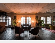 Industriell gestalteter Lounge-Bereich mit hohen, gewölbten Decken, großen Bogenfenstern und Vintage-Möbeln, darunter ein ledernes Chesterfield-Sofa, Sessel und grüne Pflanzen. Kerzenleuchter und warme Beleuchtung schaffen eine gemütliche Atmosphäre.