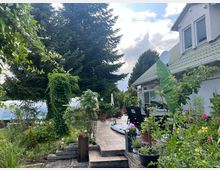Ein Garten mit gepflasterter Terrasse, umgeben von üppiger Vegetation und großen Nadelbäumen. Rechts ein modernes Wohnhaus mit grünem Ziegeldach, bodentiefen Fenstern und außenstehenden Blumentöpfen.