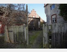 Blick durch ein geöffnetes, verwittertes Holztor in einen schmalen Hofweg zwischen alten Gebäuden aus Backstein und Putz mit roten Ziegelumrahmungen. Im Hintergrund stehen eine ziegelgedeckte Scheune und weitere Backsteinbauten, umgeben von kahlen Sträuchern und Bäumen.