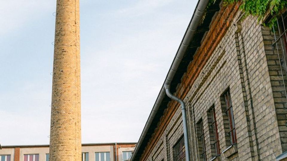 Innenhof eines alten Industriekomplexes mit hohen Backsteinfassaden, schmalen Fenstern und einem weit aufragenden Schornstein. Am rechten Gebäude sind Regenrinnen und teilweise Efeubewuchs unter dem Dach zu sehen.