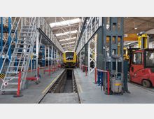 Eine Werkstatthalle mit einem gelben Zug, der auf einem Wartungsgleis steht. Die Halle enthält metallene Arbeitsplattformen, Leitern, Regale mit Materialien und Werkzeuge, sowie eine Hebebühne und ein rotes Gabelstaplerfahrzeug.