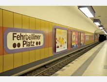 U-Bahn-Station "Fehrbelliner Platz" in Berlin mit gelb-braunen Wandverkleidungen, beleuchteten Stationsschildern und Werbeplakaten entlang des Bahnsteigs. Der Boden ist mit hellen Fliesen ausgelegt, und die Beleuchtung erfolgt durch rechteckige Deckenlampen.