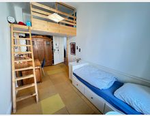 Kleines Schlafzimmer mit Hochbett aus Holz und einer Holztreppe, darunter ein Schreibtisch mit Stuhl sowie ein großer Holzkleiderschrank. Rechts befindet sich ein weißes Bett mit blauer Bettwäsche, und der gelbe Bodenbelag ergänzt das helle, minimalistische Design.