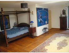 Ein Schlafzimmer mit einem Holzbettgestell im Himmelbettstil, Holzboden und einem großen, gemusterten Teppich. An der Wand hängen zwei moderne Gemälde, daneben stehen antike Möbelstücke wie ein kleiner Beistelltisch und ein Holzschrank.