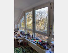 Ein lichtdurchflutetes Kinderzimmer mit einer großen Fensterfront, die den Blick auf Bäume im Herbstlaub bietet. Auf der Fensterbank und einem niedrigen Tisch sind zahlreiche Spielzeuge, darunter Modellautos, Züge und Bauklötze, sowie eine Spielmatte auf dem Boden.