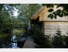 Holzhütte mit Reetdach am Ufer eines schmalen Wasserkanals, daneben ein hölzerner Steg mit einem angedockten Boot. Im Hintergrund führt eine kleine Holzbrücke über den Kanal, umgeben von dichtem Baumbestand.