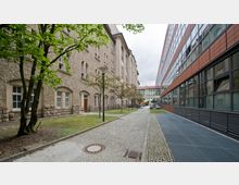 Ein schmaler gepflasterter Weg zwischen zwei Gebäuden: links ein historisches Gebäude mit beige gestrichener Fassade und mehreren Fenstern, rechts ein modernes Gebäude mit großer Glasfassade und roten Paneelen. Im Vordergrund befinden sich Bäume und Straßenlaternen.