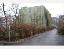 Ein moderner, begrünter Gebäude mit Kletterpflanzen bedeckt, steht an einer schmalen, asphaltierten Straße. Im Vordergrund sind kahle Büsche und ein Wegweiser, während umliegende Wohngebäude im Hintergrund sichtbar sind.