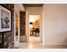 Innenraum eines Gebäudes mit moderner Einrichtung und rustikalem Stil, gekennzeichnet durch Wände aus freiliegendem Ziegelstein. Im Vordergrund ist ein Flur mit Fliesenboden, links eine Ziegelwand mit einem gerahmten Foto, und im Hintergrund ein Arbeitsbereich mit einem Schreibtisch, einer Pflanze und einer Stehlampe.